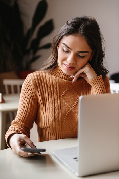 ritratto di una giovane donna abbastanza bella seduta in un caffè al chiuso utilizzando il computer portatile e il telefono cellulare.