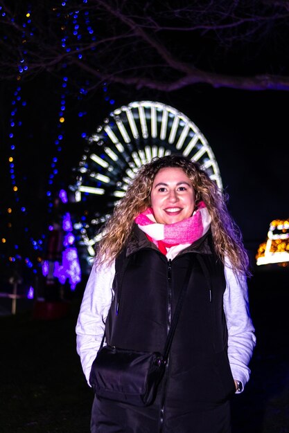 Ritratto di una giovane donna adulta che sorride guardando la macchina fotografica in un parco di Natale di notte Sullo sfondo una ruota panoramica e alberi decorati con luci di Natale