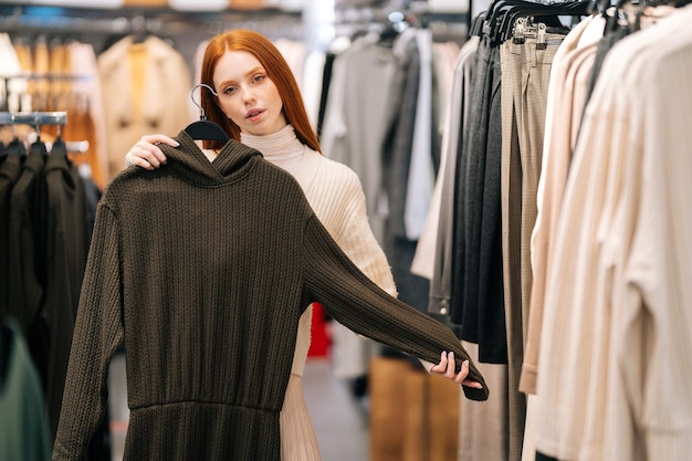 Ritratto di una giovane donna affascinante che sceglie i vestiti e cerca di specchiarsi nel negozio di abbigliamento