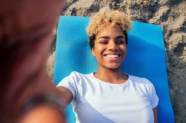 Ritratto di una giovane donna africana latina sdraiata su un tappetino da yoga in spiaggia, scatta foto selfie sullo smartphone della fotocamera vista dall'alto