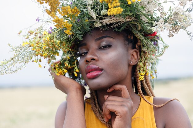 Ritratto di una giovane donna afroamericana, modella di moda, con grandi fiori tra i capelli. Ritratto di una ragazza in un piano storto in un campo con fiori. corona sulla sua testa on