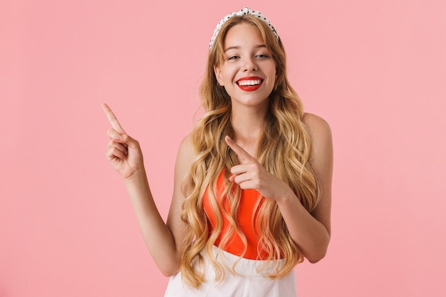 Ritratto di una giovane donna allegra con lunghi capelli ricci che sorride e punta le dita al copyspace isolato su un muro rosa
