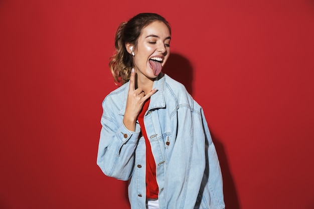 Ritratto di una giovane donna allegra ed elegante che indossa una giacca di jeans in piedi isolata sul muro rosso, ascoltando musica con auricolari wireless, cantando