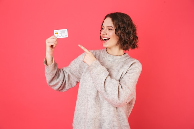 Ritratto di una giovane donna allegra in piedi isolato su rosa, presentando la carta di credito in plastica