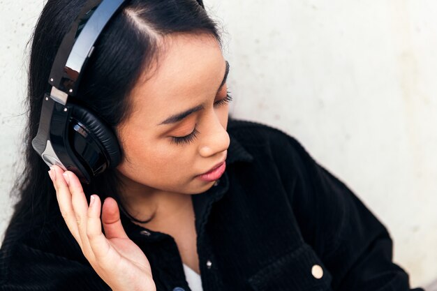 Ritratto di una giovane donna asiatica con le cuffie che si diverte ad ascoltare musica all'aperto appoggiata a un muro di cemento