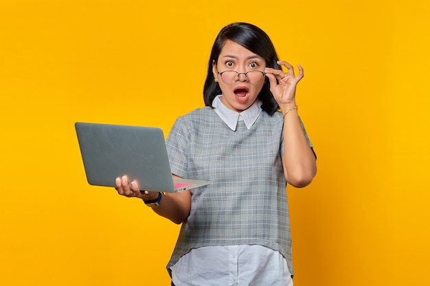 Ritratto di una giovane donna asiatica sorpresa che tiene in mano un laptop e tocca gli occhiali con un dito su sfondo giallo