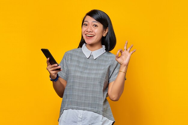 Ritratto di una giovane donna asiatica sorridente che mostra il segno giusto e tiene in mano uno smartphone su sfondo giallo