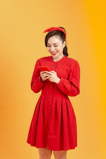 Ritratto di una giovane donna asiatica sorridente in abito rosso che tiene in mano uno smartphone su sfondo arancione
