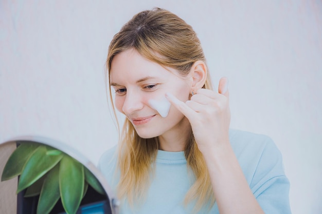 Ritratto di una giovane donna attraente che applica una crema idratante sul viso a casa Pulizia della pelle con schiuma e idratazione della pelle con cosmetologia in crema e concetto di cura della pelle a casa