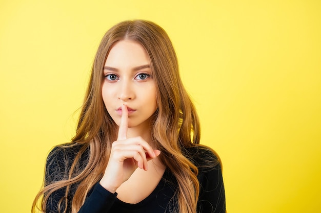 Ritratto di una giovane donna attraente con capelli lunghi e trucco tiene un dito vicino alle labbra in studio su uno sfondo giallo. il concetto di segretezza e silenzio.