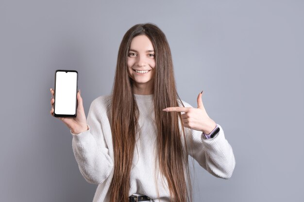 Ritratto di una giovane donna attraente ragazza con i capelli lunghi scuri che sorride facilmente e punta il dito contro uno smartphone cellulare vuoto su uno sfondo grigio studio. Posto per il testo. Con copia spazio.