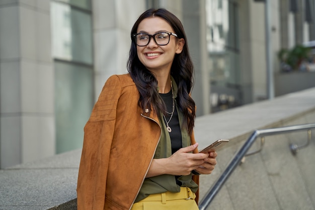 Ritratto di una giovane donna bella e alla moda con gli occhiali che tiene il suo smartphone