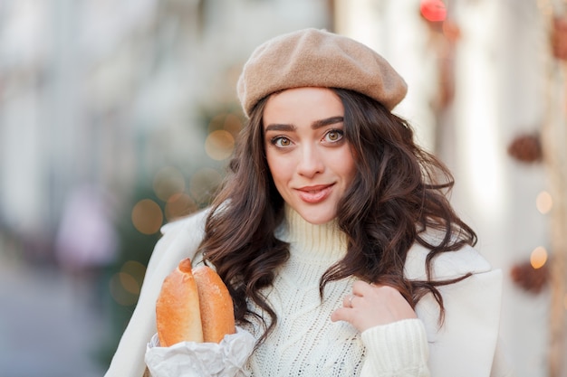 Ritratto di una giovane donna bellissima in un berretto in una città europea. La giovane donna tiene un sacchetto di carta con le baguette. Natale. Vacanze.