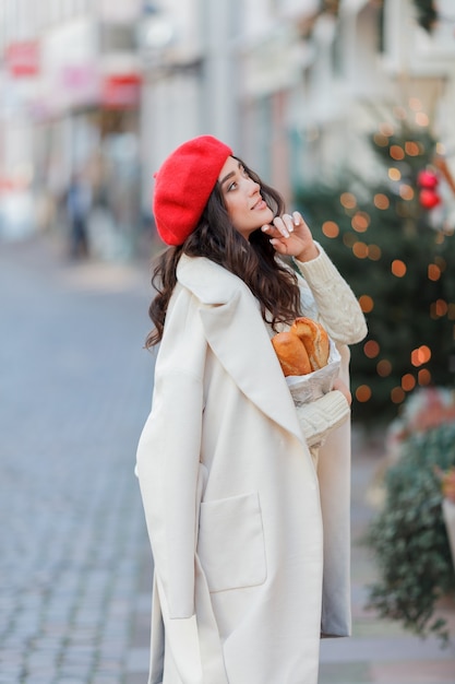 Ritratto di una giovane donna bellissima in un berretto rosso in una città europea. La giovane donna tiene un sacchetto di carta con le baguette. Natale. Vacanze.