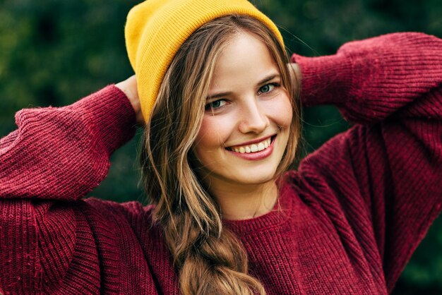 Ritratto di una giovane donna bionda che sorride ampiamente con un sano sorriso a trentadue denti che indossa un maglione rosso e un cappello giallo in posa sullo sfondo della natura nel parco La gente viaggia lo stile di vita