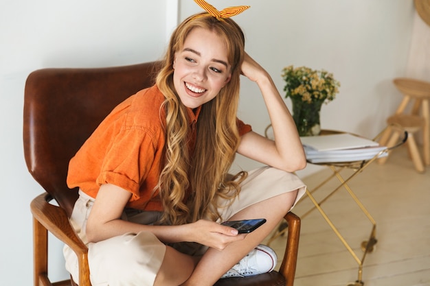 Ritratto di una giovane donna bionda positiva carina a casa al chiuso utilizzando il telefono cellulare seduto sulla sedia.