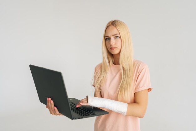 Ritratto di una giovane donna bionda seria con il braccio rotto avvolto in una benda di gesso che tiene il computer portatile guardando la fotocamera in piedi su sfondo bianco isolato