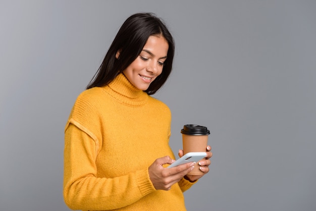 Ritratto di una giovane donna casual felice isolata sopra il muro grigio, tenendo il caffè da asporto, utilizzando il telefono cellulare