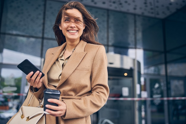 Ritratto di una giovane donna caucasica allegra sorridente con uno smartphone e un caffè in piedi fuori