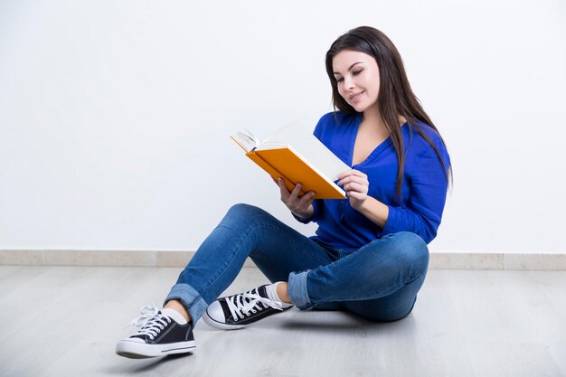 Ritratto di una giovane donna che indossa blue jeans e una camicetta e seduta sul pavimento con un libro arancione e leggendolo.