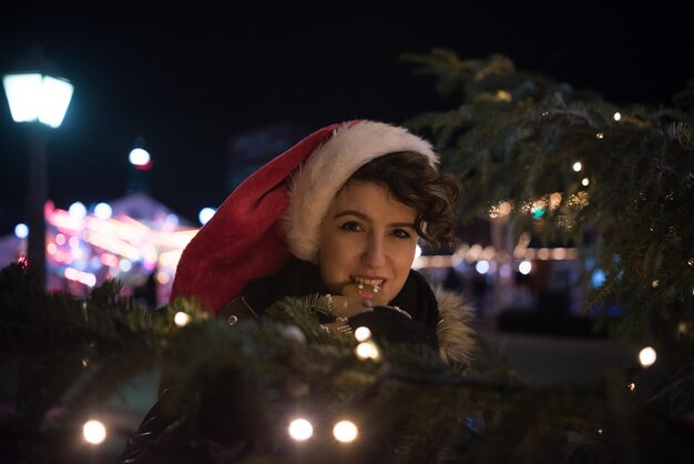 Ritratto di una giovane donna che mangia un biscotto vicino all'albero di Natale illuminato di notte