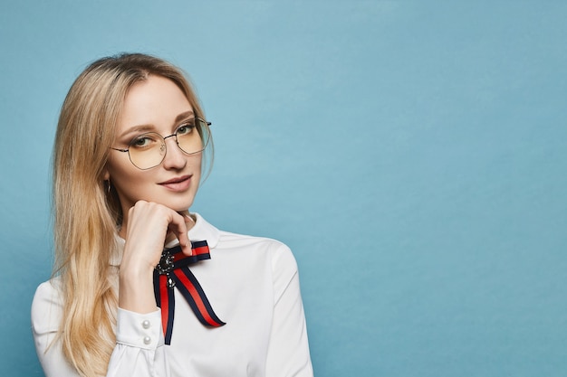 Ritratto di una giovane donna con i capelli biondi in camicia bianca e occhiali, isolato su sfondo azzurro con spazio di copia. Ragazza astuta dello studente in blusa bianca e vetri che esaminano la macchina fotografica