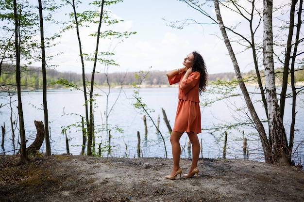 Ritratto di una giovane donna con un sorriso sul viso, emozioni felici. Una ragazza con lunghi capelli ricci ondulati in un vestito in estate sulla natura, in una foresta su un lago, stava vicino a una strada e a cespugli