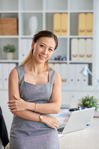 Ritratto di una giovane donna d'affari asiatica sorridente in abito formale che tiene il gomito dell'altro braccio mentre si appoggia al tavolo dell'ufficio con il laptop