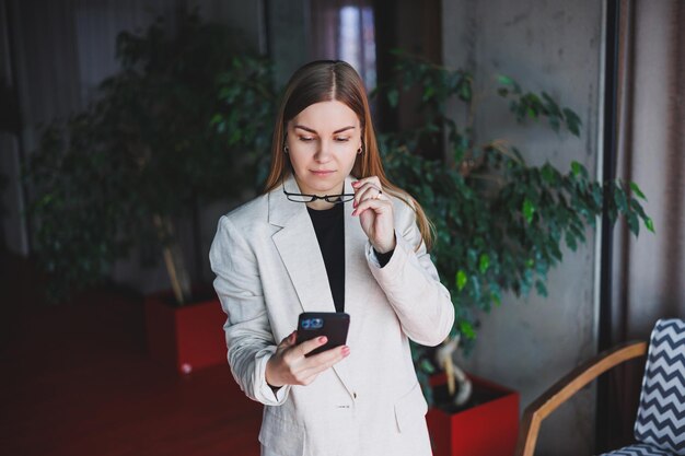 Ritratto di una giovane donna dai capelli biondi con una giacca beige e occhiali sul viso un telefono mod nelle mani di una donna Bella ragazza blogger moderna