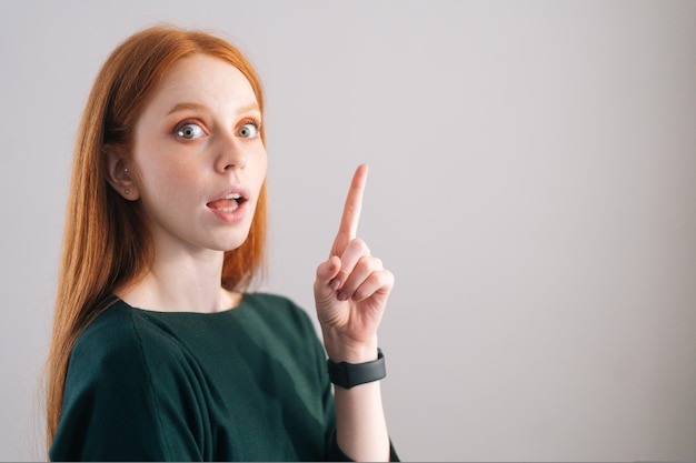 Ritratto di una giovane donna dai capelli rossi stupita che punta verso l'alto con il dito guardando la telecamera su bianco