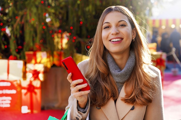 Ritratto di una giovane donna eccitata che tiene in mano uno smartphone e guarda la telecamera con un albero di Natale e regali sullo sfondo all'aperto