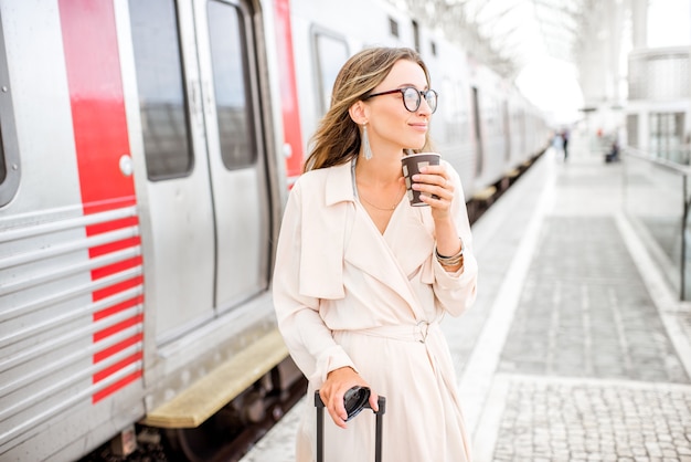 Ritratto di una giovane donna elegante in piedi con una tazza di caffè vicino al treno alla stazione ferroviaria