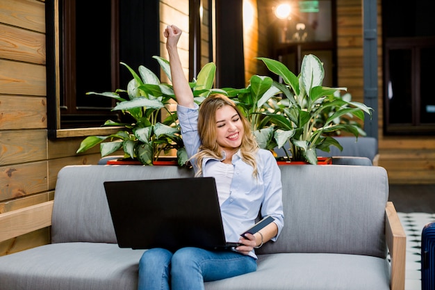 Ritratto di una giovane donna emozionante nell'abbigliamento casual che si siede sul sofà nel posto coworking moderno del sottotetto