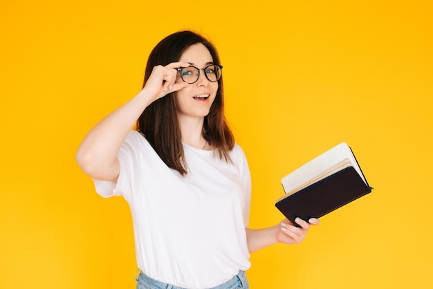 Ritratto di una giovane donna felice che indossa occhiali e maglietta bianca immersa nella lettura di un libro con a