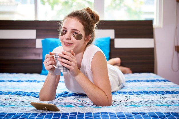 Ritratto di una giovane donna felice che riposa sul letto, beve una tazza di tè caldo e guarda l'episodio dello spettacolo sullo schermo dello smartphone