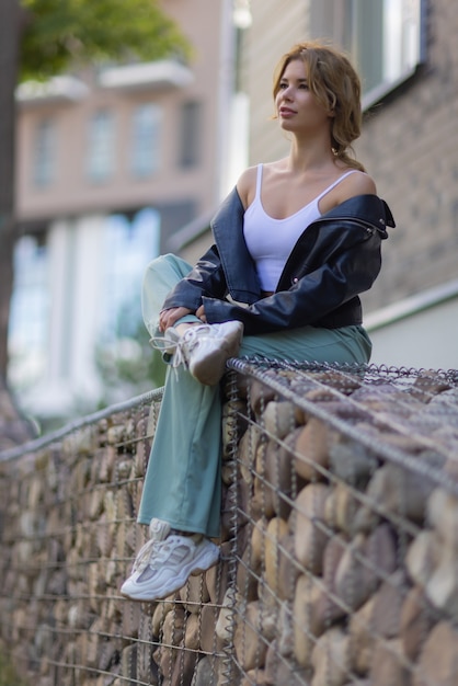 Ritratto di una giovane donna graziosa in pantaloncini blu seduta su una panchina nel cortile di un complesso residenziale.