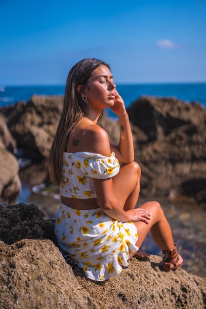 Ritratto di una giovane donna in abito bianco sulla spiaggia seduta su alcune rocce ad ascoltare il suono delle onde e del mare