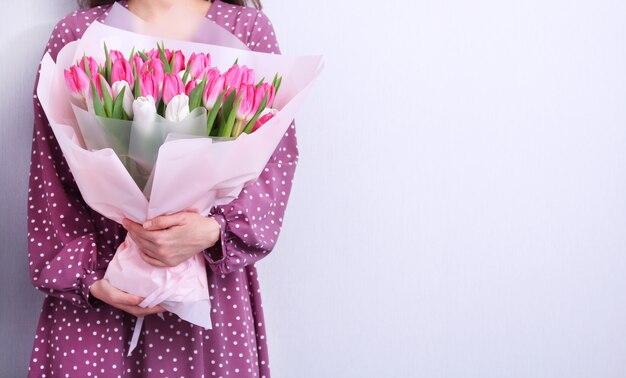 Ritratto di una giovane donna in abito viola azienda bouquet di tulipani rosa e bianchi su grigio. Cartolina d'auguri di primavera. Pasqua, concetto di fiore di primavera. Festa della mamma o della donna.