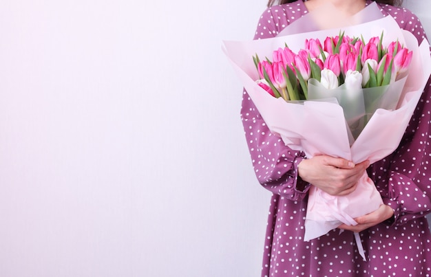 Ritratto di una giovane donna in abito viola azienda bouquet di tulipani rosa e bianchi su grigio. Cartolina d'auguri di primavera. Pasqua, concetto di fiore di primavera. Festa della mamma o della donna.