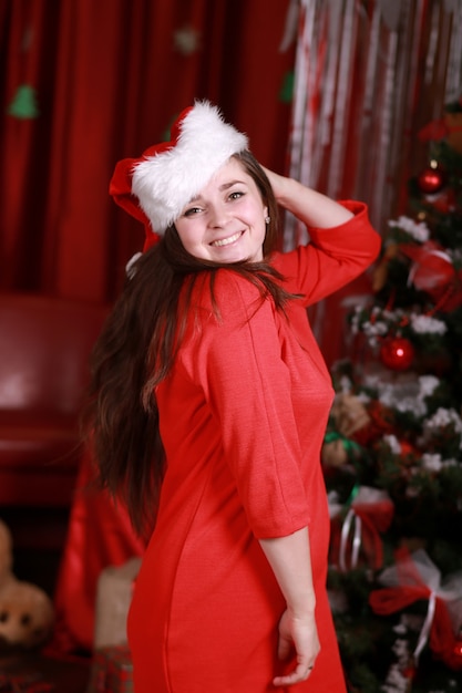 Ritratto di una giovane donna in santa helper cappello e abito rosso in preparazione per il Natale a casa. natale, natale, inverno, concetto di felicità - donna sorridente sul muro dell'albero di Natale.