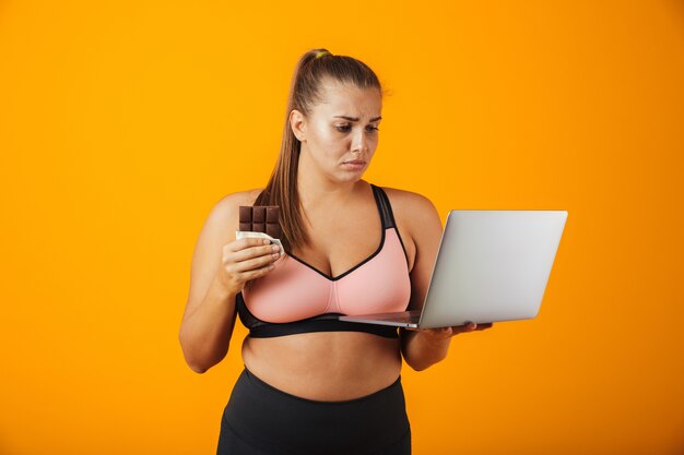 Ritratto di una giovane donna in sovrappeso delusa che indossa abbigliamento sportivo in piedi isolato sopra la parete gialla, utilizzando il computer portatile, tenendo la barretta di cioccolato