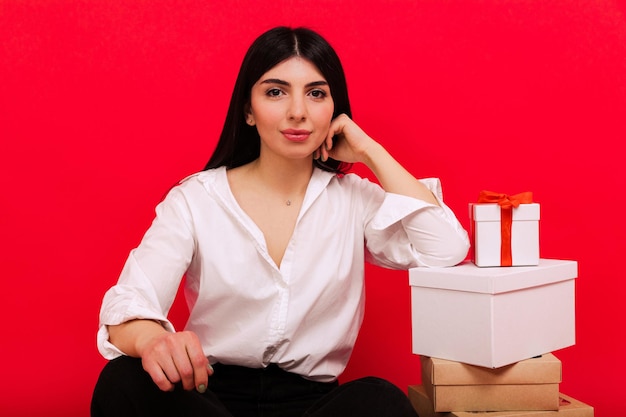 Ritratto di una giovane donna in una camicia bianca e jeans seduta accanto ai regali di Natale su sfondo rosso