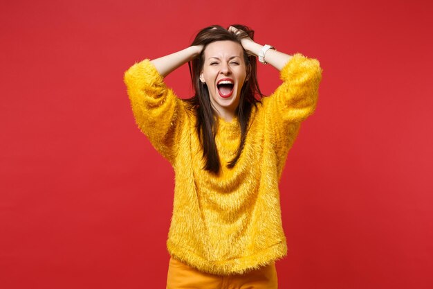 Ritratto di una giovane donna pazza e divertente in maglione di pelliccia gialla che urla, aggrappandosi alla testa isolata su sfondo rosso brillante della parete in studio. Persone sincere emozioni, concetto di stile di vita. Mock up copia spazio.