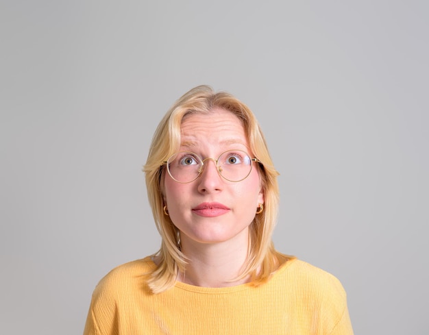 Ritratto di una giovane donna pensierosa con i capelli biondi e gli occhiali che guarda su uno sfondo bianco