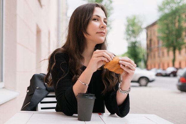 Ritratto di una giovane donna seduta in un caffè estivo e mangiare un croissant con caffè Buon umore e riposo in estate