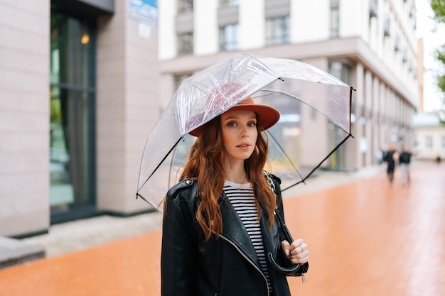 Ritratto di una giovane donna seria allo zenzero che indossa un cappello elegante in piedi con un ombrello trasparente sulla strada in caso di pioggia che guarda l'obbiettivo