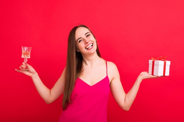 Ritratto di una giovane donna sorridente felice in un abito elegante rosa con in mano un bicchiere di champagne e una scatola con un regaloFesta di festa