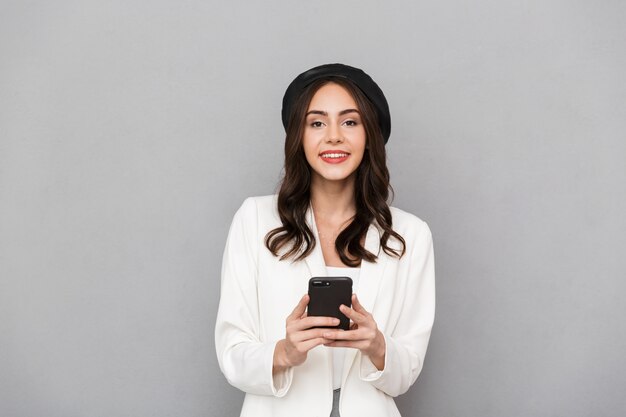 Ritratto di una giovane donna sorridente vestita in giacca su sfondo grigio, utilizzando il telefono cellulare