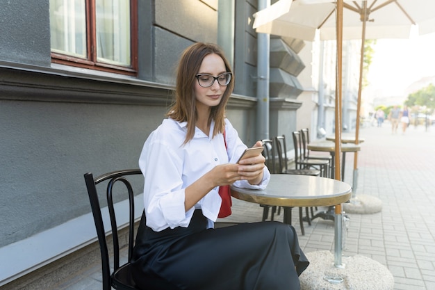 Ritratto di una giovane donna sulla terrazza estiva di un caffè e digitando sul telefono