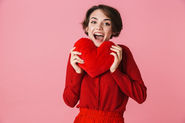 Ritratto di una giovane e bella donna che indossa un abito rosso in piedi isolato su sfondo rosa, mostrando il cuscino a forma di cuore